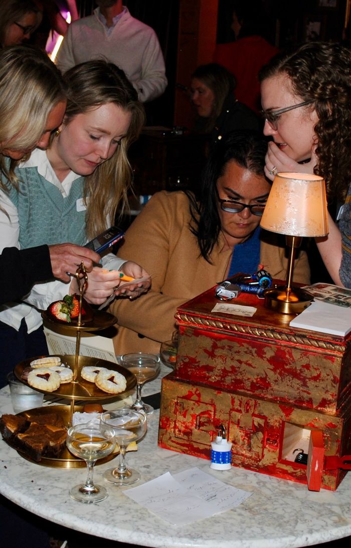 Guests gathered around a locked red and gold puzzle box, working out clues at Révéler